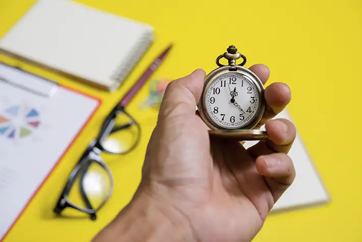 Mão segurando um relógio de bolso antigo sobre fundo amarelo com materiais de escritório, ilustrando o conceito de reduzir Lead Time e gestão de tempo em processos Lean.