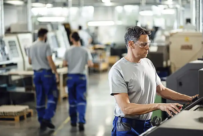 Operador de perfil, usando óculos de proteção e camiseta cinza, manuseando o painel de controle de uma máquina CNC em uma fábrica moderna e iluminada. Ao fundo, outros trabalhadores de uniforme azul caminham pelo galpão industrial.